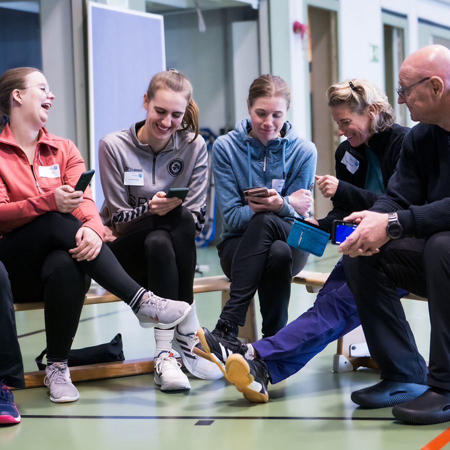 5 Personen sitzen in Sporthalle und schauen auf ihre Handys