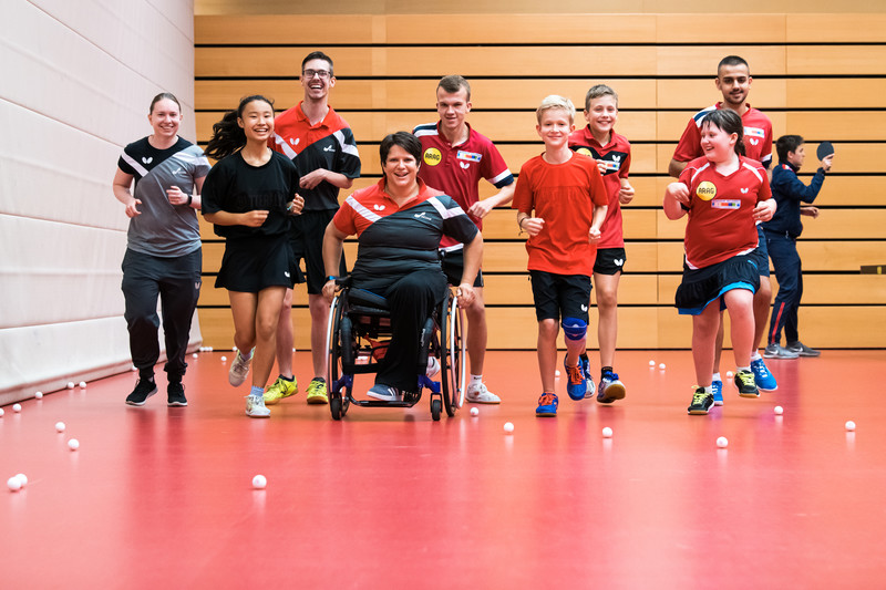 Gruppe von Kindern und Jugendlichen, darunter ein Rollstuhlfahrer, beim gemeinsamen Sport in einer Halle mit Tischtennisbällen auf dem Boden.