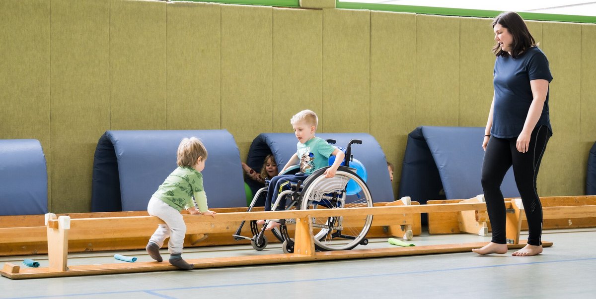 Ein Kind im Rollstuhl und ein Kleinkind spielen gemeinsam auf einer Turnbank in einer Sporthalle
