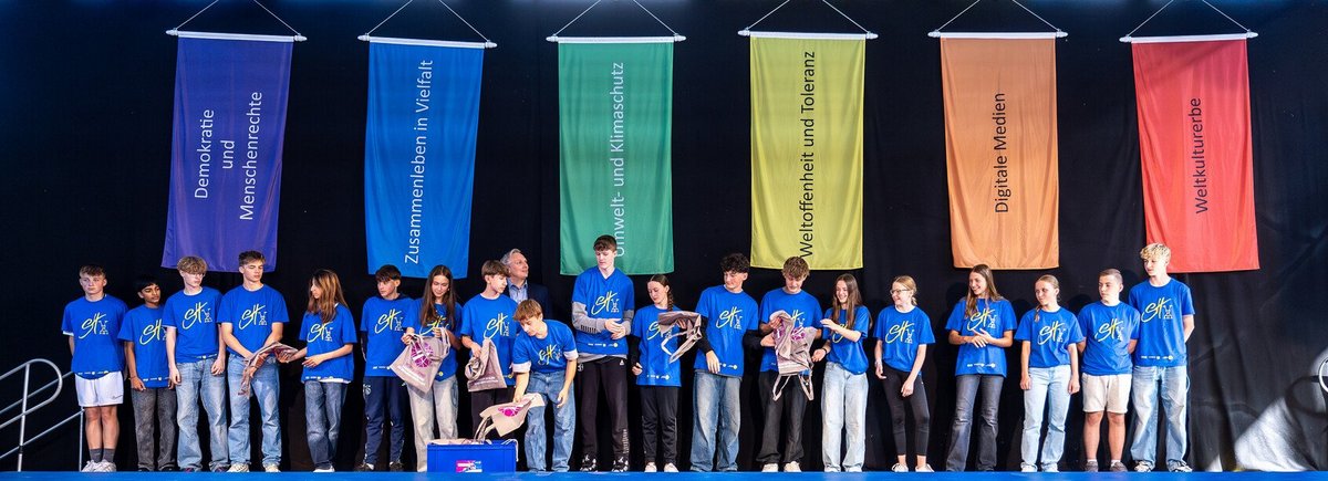 Auf einer Bühne steht eine Gruppe Sporthelfer*innen, im Hintergrund hängen Banner mit Werten im Sport, wie z. B. Demokratie, Vielfalt und Toleranz