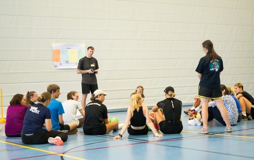Menschen sitzen in einer Sporthalle im Kreis, um die Übungsleitung herum