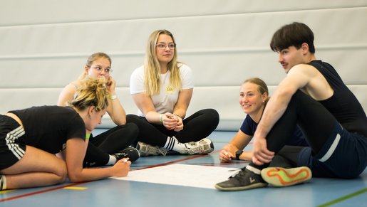 Junge Erwachsene sitzen im Kreis in einer Sporthalle