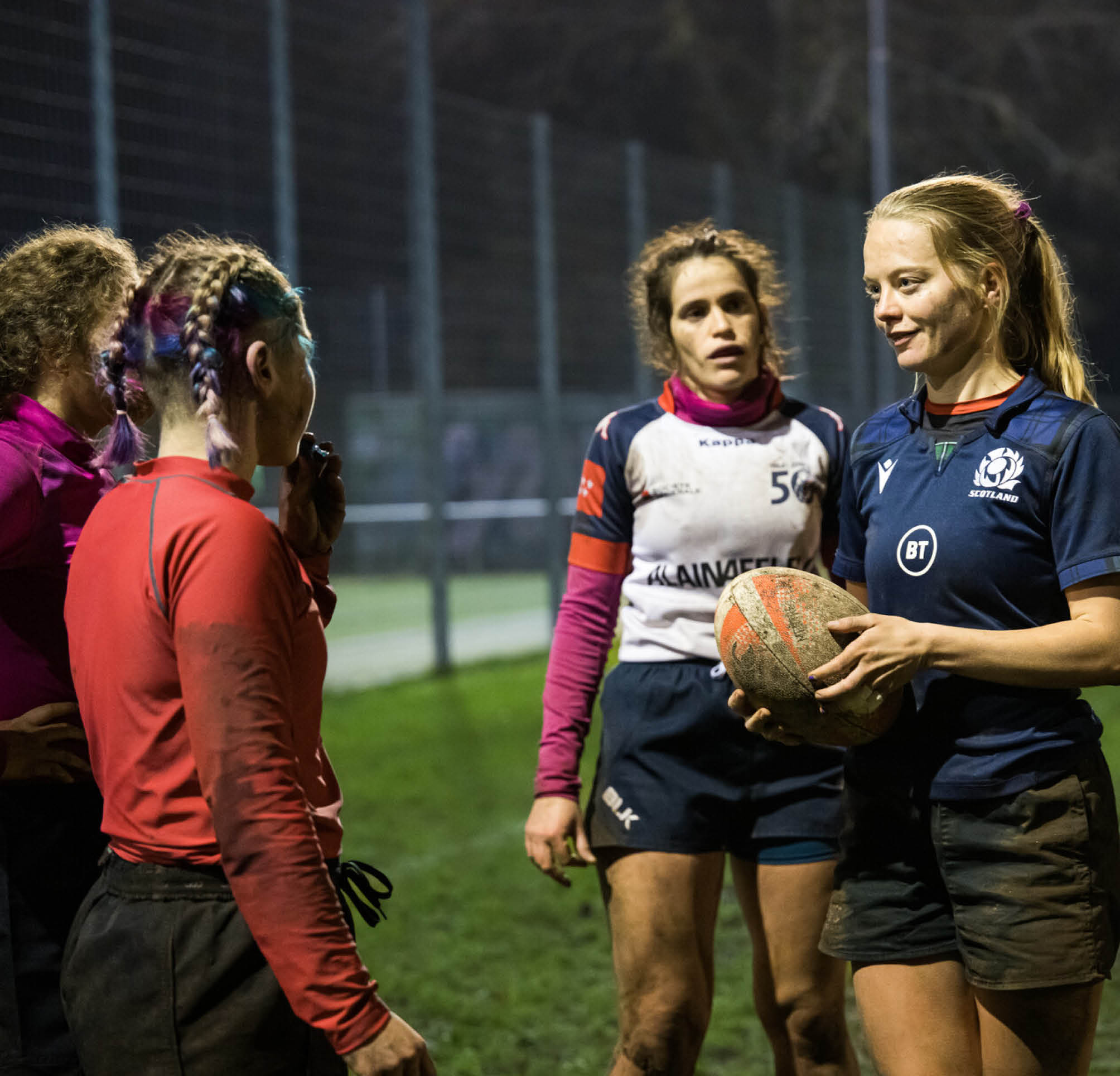 Foto mehrerer Rugby-Spielerinnen die Abends bei Flutlicht auf einem gr nen Rasen stehen. Die rechte Spielerin h lt einen Rugbyball in der Hand